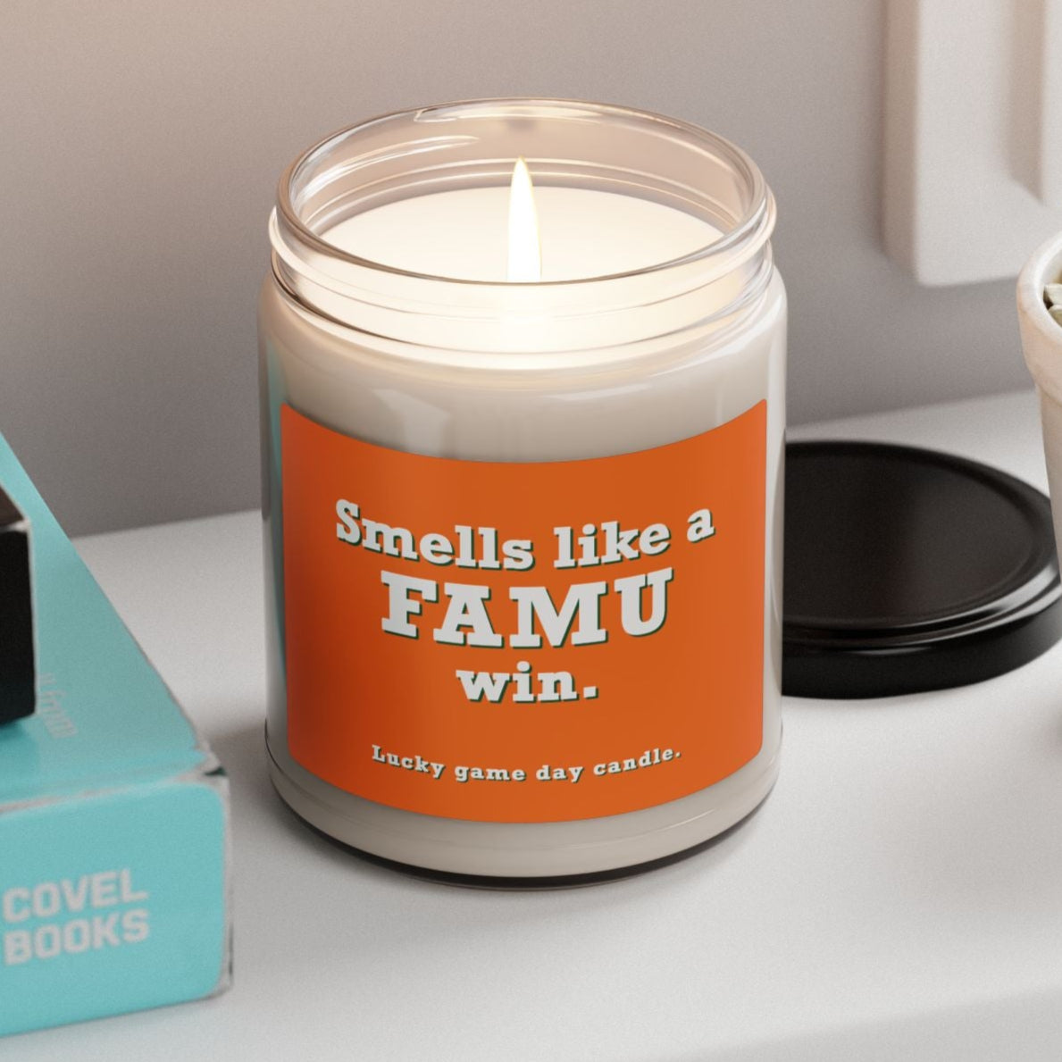 Candle with 'Smells like a FAMU win.' label on a shelf with books and a plant