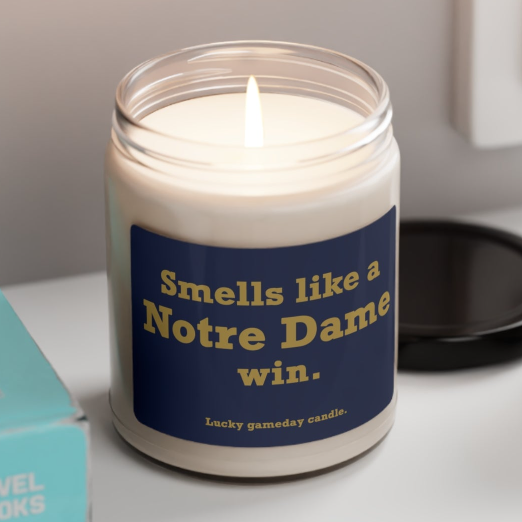 A clear glass jar candle with a white label that reads 'SMELLS LIKE A NOTRE DAME WIN.' The candle is placed on a table with a box in the background, indicating it might be a gift or a decorative item for a fan.