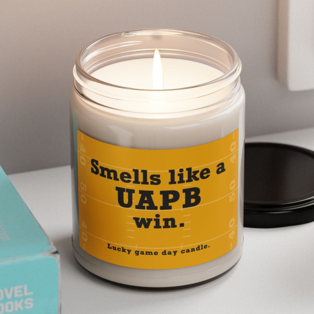 Arkansas Pine-Bluff candle with a yellow label on a shelf next to books and a plant